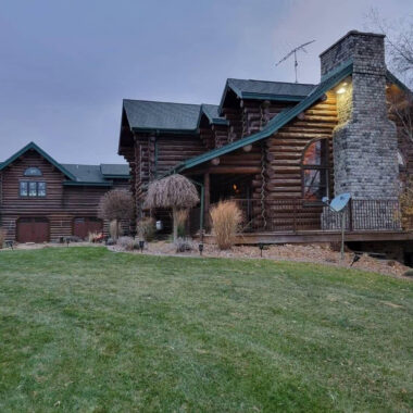 Log cabin-style house with a stone chimney, green roof, and front deck overlooking a grassy yard at dusk.