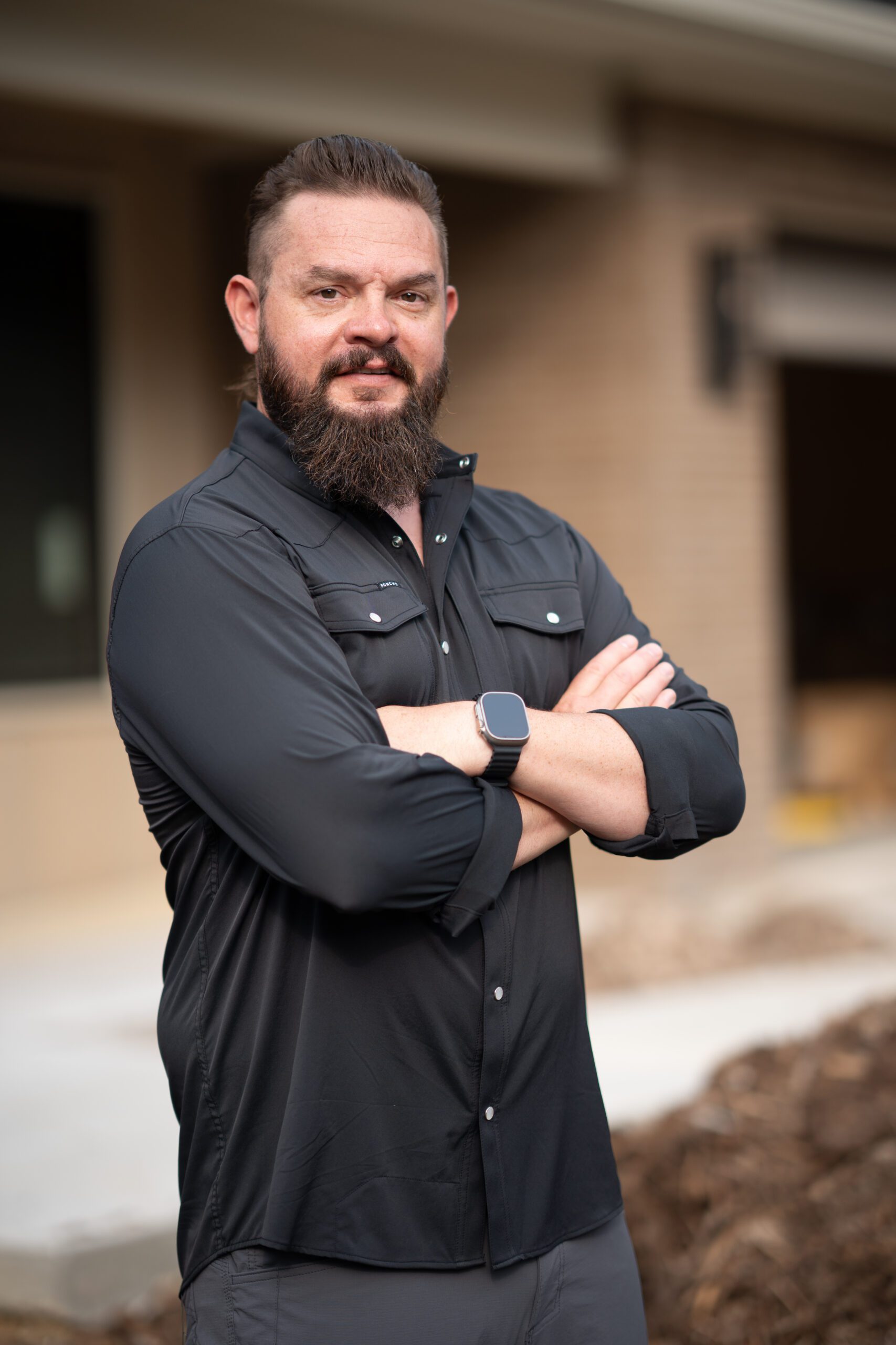 Bearded man in a black shirt standing outdoors with arms crossed in front of a building exterior.