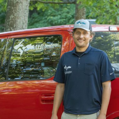 Smiling man in a navy polo shirt stands beside a red U.P. Roofing Company truck outdoors with trees in the background.